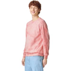 A pink crewneck sweatshirt displayed on a model, featuring a subtle tie-dye effect and a logo on the sleeve.