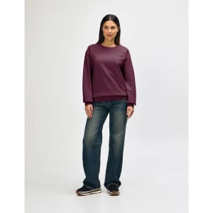 A woman stands wearing a burgundy Gildan Heavy Blend adult crewneck sweatshirt and blue jeans. The sweatshirt features a logo.