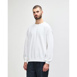 A white crewneck sweatshirt worn by a model, featuring long sleeves and a relaxed fit. It has a logo on it.