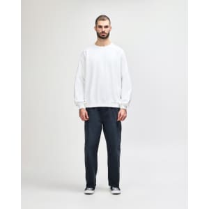 A white crewneck sweatshirt paired with black pants, worn by a model walking. The sweatshirt features a logo.