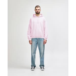 A man stands wearing a pink hooded sweatshirt and black jeans, with his hands in the pockets of the sweatshirt.