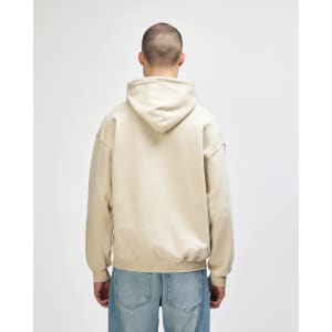 A beige hooded sweatshirt with a back view, featuring a hood and casual fit. The model wears blue jeans.