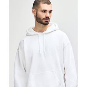 A white Gildan Heavy Blend adult hooded sweatshirt is worn by a young man adjusting the hood. The product features a logo.