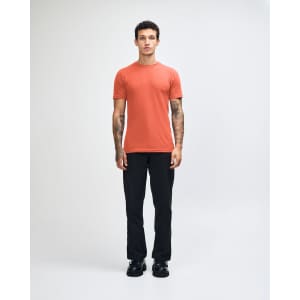 A model wearing an orange unisex short sleeve t-shirt and beige pants, standing casually. The t-shirt has a logo.