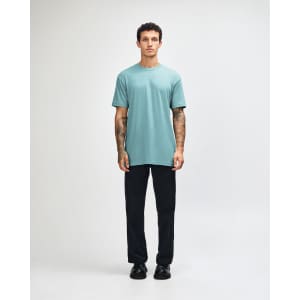 A unisex short sleeve t-shirt in a teal colour, paired with black cargo pants and casual shoes. The model has a relaxed pose.