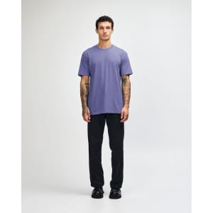 A unisex short sleeve t-shirt in purple, worn by a model with black pants and sneakers, featuring a logo.
