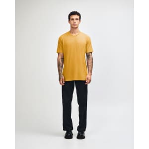 A unisex short sleeve t-shirt in mustard yellow, worn with black cargo pants, featuring a logo.