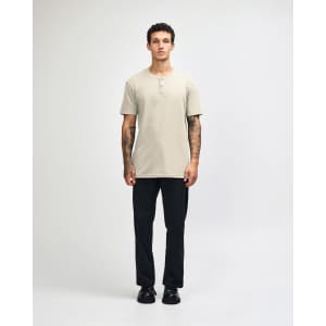 A beige unisex Henley T-shirt featured on a model paired with light blue jeans and black sneakers. The shirt has a logo.