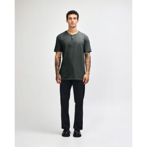 A man stands wearing a dark grey unisex Henley T-shirt paired with light blue jeans and black sneakers.