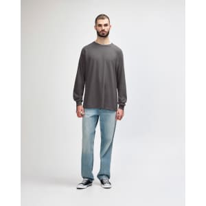 A long-sleeve T-shirt in grey worn by a model, paired with black jeans and sneakers. The shirt features a logo.