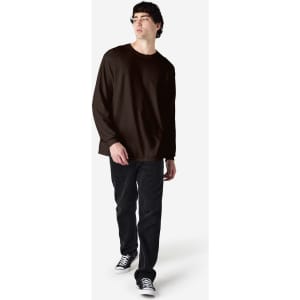 A man wearing a brown long sleeve T-shirt and black jeans, standing against a plain background.