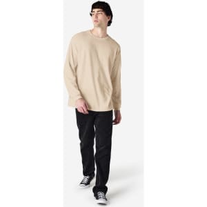 A man is wearing a beige long sleeve T-shirt paired with black jeans and sneakers, featuring a logo.