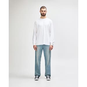 A white long sleeve T-shirt worn by a man, paired with black pants and shoes, standing against a light background.