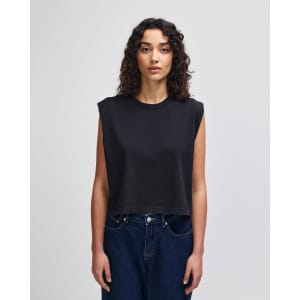 A women's muscle t-shirt in dark grey, featuring a cropped, sleeveless design. The model has curly hair and wears jeans.