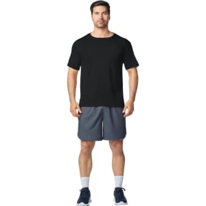 A man stands wearing a black Gildan Performance adult T-shirt and grey shorts, with sports shoes and socks.