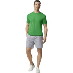 A man wearing a green Gildan Performance adult t-shirt and grey shorts, standing casually. The shirt features a logo.