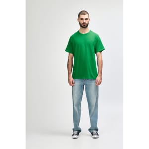 A green Gildan Heavy Cotton adult t-shirt displayed on a model wearing black jeans and sneakers, with a logo present.