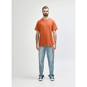 An adult wearing an orange Gildan Heavy Cotton T-shirt, paired with black pants and black shoes. The shirt has a logo.