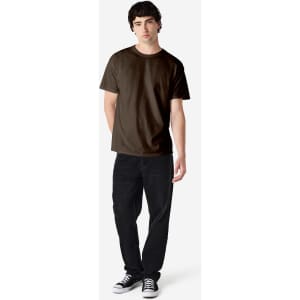 A man stands wearing a dark brown Gildan Heavy Cotton adult t-shirt and black jeans, with a logo visible on the shirt.