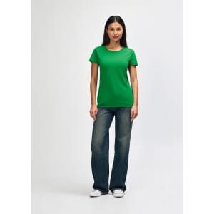 A woman stands wearing a green Gildan Heavy Cotton Ladies' T-Shirt and blue jeans, with a casual pose and white shoes.