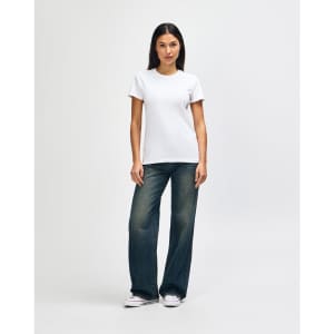 A woman stands in a white Gildan Heavy Cotton Ladies' T-Shirt paired with blue jeans and white sneakers.