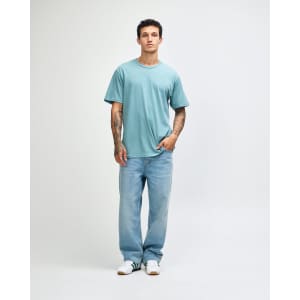 A man wearing a light blue unisex short sleeve t-shirt with denim jeans, standing against a plain background.