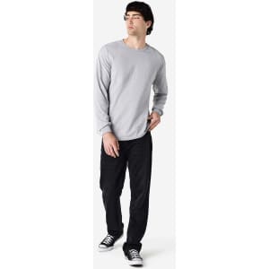 A model wearing a grey long sleeve t-shirt paired with black trousers and sneakers, standing against a plain background.