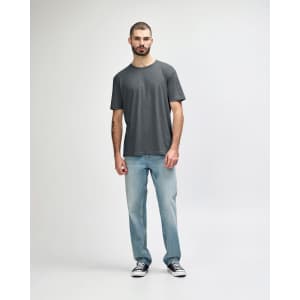 A man stands wearing a grey Gildan Softstyle adult t-shirt paired with light blue jeans and white shoes.
