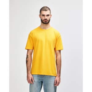 A person stands wearing a bright orange Gildan Softstyle adult T-shirt, paired with grey jeans. The shirt has a logo.