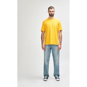 A man stands wearing a bright yellow Gildan Softstyle adult T-shirt paired with grey jeans. The shirt has a logo.