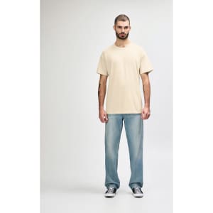 A model stands wearing a cream Gildan Softstyle adult T-shirt paired with light blue jeans and white sneakers.