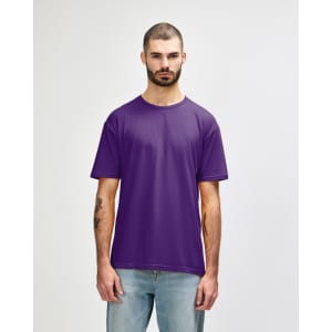 A man stands wearing a purple Gildan Softstyle adult T-shirt, paired with light blue jeans. The shirt has a logo.