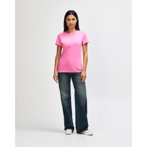 A pink Gildan Softstyle ladies' t-shirt paired with blue jeans and white sneakers, featuring a logo.