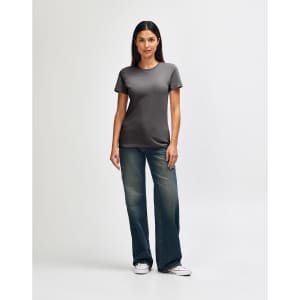 A woman stands wearing a soft-style ladies' t-shirt in grey, paired with blue jeans and white sneakers.