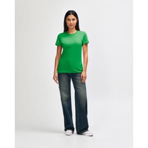 A green Gildan Softstyle ladies' t-shirt paired with blue jeans and white sneakers, modelled by a woman.