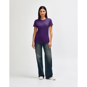 A woman is wearing a purple Gildan Softstyle ladies' T-shirt paired with blue jeans and white sneakers.