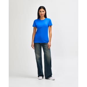 A woman stands wearing a blue Gildan Softstyle ladies' t-shirt and denim pants, paired with white sneakers.