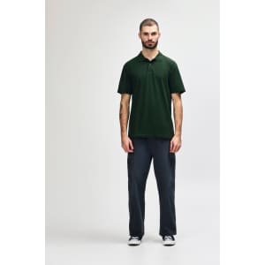 A man stands wearing a green Gildan Softstyle adult double pique polo, paired with black pants and shoes.