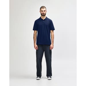A man stands wearing a navy blue Gildan Softstyle adult double pique polo and black jeans. The polo features a logo.