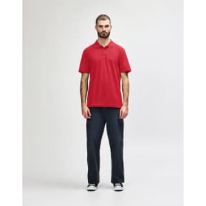 A red Gildan Softstyle adult double pique polo shirt displayed on a model wearing black jeans and sneakers. The shirt has a logo.