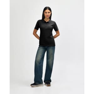 A woman poses in a black Gildan Softstyle Ladies' Double Pique Polo, paired with blue jeans and white sneakers.