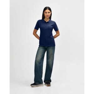 A woman stands smiling in a navy blue Gildan Softstyle ladies' polo shirt and denim jeans, wearing white sneakers.
