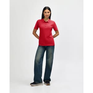 A model wearing a red Gildan Softstyle ladies' double pique polo with jeans and white sneakers. The image features a logo.