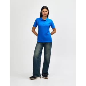 A woman poses in a bright blue polo shirt and jeans, standing confidently with arms crossed. The polo features a logo.