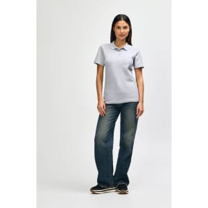 A woman wearing a grey Gildan Softstyle ladies' double pique polo and blue jeans, standing casually.
