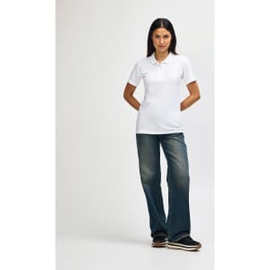 A model wearing a white Gildan Softstyle Ladies' Double Pique Polo with blue jeans and white sneakers, featuring a logo.