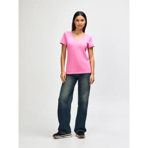 A woman stands wearing a pink Gildan Softstyle ladies' V-neck T-shirt and blue jeans, with white sneakers.