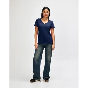 A woman is wearing a navy Gildan Softstyle ladies' V-neck T-shirt, paired with blue jeans and white sneakers.