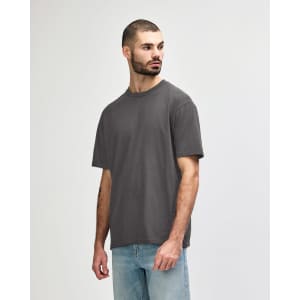 A man stands wearing a midweight T-shirt in olive green. The shirt features a logo on it.