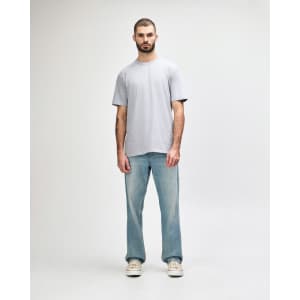 A man stands wearing a light grey Gildan Softstyle adult midweight T-shirt and blue jeans. The shirt has a logo.